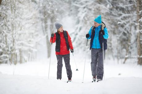Active senior couple on skis moving down snowdrift in snowfall during training in winter forest