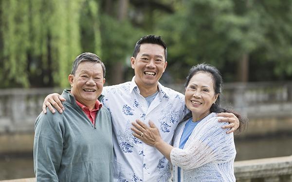 Portrait of an Asian man standing in the park with his parents. The son is in the middle, with his arms around his parents shoulders. They are looking at the camera, smiling.