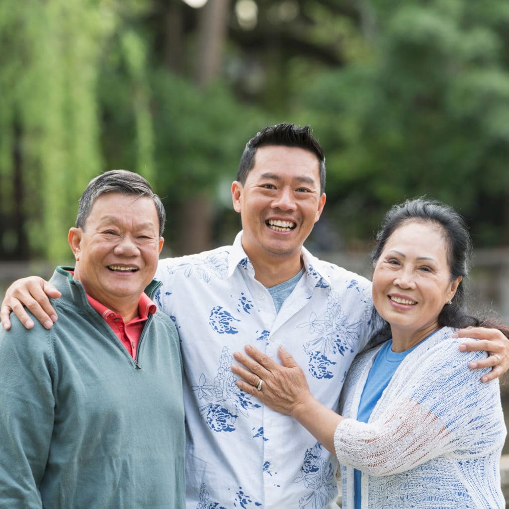 Portrait of an Asian man standing in the park with his parents. The son is in the middle, with his arms around his parents shoulders. They are looking at the camera, smiling.