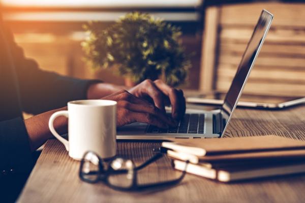 Close up of a person typing on a laptop with a mug, pair of glasses and notebooks on the desk