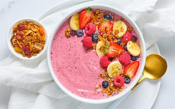 Smoothie Bowl topped with bananas, strawberries, blueberries and granola on top of a white plate with a gold spoon beside it